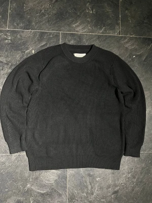 Svart stickad tröja Maison Margiela - Säljer en svart, ribbstickad tröja från Maison Margiela. Klassisk rund halsringning och långa ärmar. Tröjan har de ikoniska vita sömmarna i nacken och är perfekt för dig som gillar minimalistisk och stilren design.