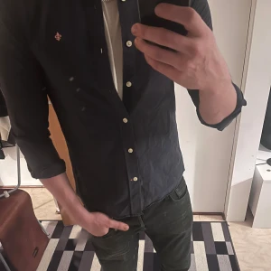 Mörkblå skjorta från Morris - Säljer en stilren mörkblå skjorta från Morris med klassisk krage och vita knappar. Skjortan har lång ärm och en snygg broderad logga på bröstet. Perfekt för dig som vill ha en clean look med lite preppy vibe.