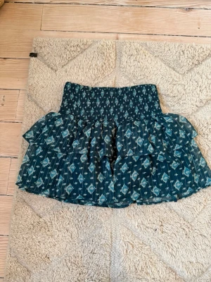 Mönstrad volangkjol från Brownie - Supersöt kort kjol från Brownie i mörkblått med vitt mönster. Inbyggda shorts!! Kjolen har bred smock i midjan och dubbla volanger som ger en luftig och flowy känsla. Perfekt till linne eller crop top. Materialet är lätt och känns som polyester.