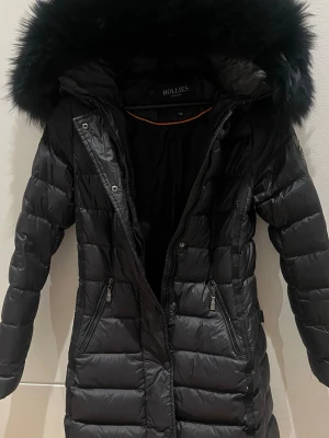 Svart lång pufferjacka Hollies - Säljer en svart lång pufferjacka från Hollies med stor fluffig huva och dragkedja framtill. Jackan har quiltad design, två fickor med dragkedja och snygg pälskant på huvan. Perfekt för kalla dagar och riktigt varm.