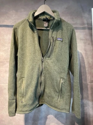 Patagonia fleece  - Mysig grön fleecekofta från Patagonia med dragkedja framtill och praktiska fickor med dragkedja. Perfekt för kyliga dagar och outdoor-häng. Klassisk logga på bröstet och hög krage för extra värme.