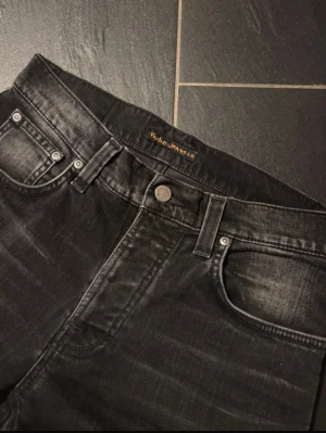 Svarta slim fit jeans från Nudie Jeans - Snygga svarta jeans från Nudie Jeans med slim passform och klassisk femficksdesign. Jeansen har slitna detaljer och subtila tvättade partier som ger en cool look. Materialet är jeans och bakfickorna har Nudie Jeans signaturbroderi.