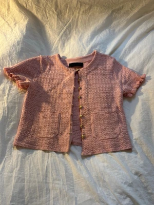 Rosa kofta från Zara med guldknappar - Supersöt rosa kofta från Zara med korta ärmar och volangkant. Koftan har dekorativa guldknappar framtill, två fickor och ett strukturerat tyg som känns lyxigt. Perfekt för vår och sommar när du vill ha en chic look.