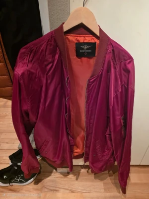 Vinröd bomberjacka från Assteries - Säljer en vinröd bomberjacka från Assteries med orange innerfoder. Jackan har dragkedja framtill, fickor på sidorna och en klassisk ärmficka. Materialet är glansigt och känns lätt, perfekt för dig som vill ha en snygg och enkel jacka.