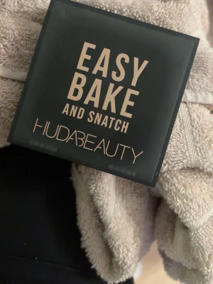 Huda Beauty Easy Bake Cupcake puder - Pressed brightening och setting powder från Huda Beauty i färgen Cupcake. Kompakt puder i en svart fyrkantig dosa med spegel, perfekt för att ljusa upp och fixera sminket. Pudret är ljusbeige och har en silkeslen finish. Använt max 10 ggr