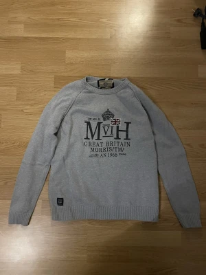 Grå stickad tröja från Morris - Snygg grå stickad tröja från Morris med tryck och brittisk flagga på bröstet. Tröjan har ribbade muddar vid ärmslut och nederkant samt lång ärm. Perfekt för dig som gillar klassisk stil med en modern twist.