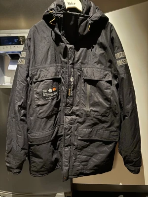 Svart Sail Racing Glacier Bay Parka - Svart parkajacka från Sail Racing, modell Glacier Bay Parka. Jackan har huva, flera stora fickor med lock och dragkedja, samt coola detaljer och tryck på ärmen och bröstet. Perfekt för kalla dagar och har en riktigt schysst streetstil.
