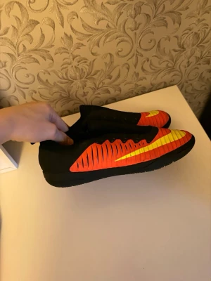 Nike innomhusskor  - Säljer ett par grymma Nike fotbollsskor i svart med röda och gula detaljer. Skorna har en låg profil, platt sula och ett coolt mönster på sidan. Perfekta för dig som vill sticka ut på planen och få bra grepp inomhus.
