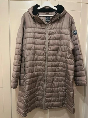 Ljusbrun pufferjacka från NorthWest - Säljer en lång, ljusbrun pufferjacka från NorthWest med huva och hel dragkedja. Jackan har quiltad design, svart fleecefoder i huvan och en badge på ena ärmen. Perfekt för kyliga dagar när du vill vara både varm och snygg.