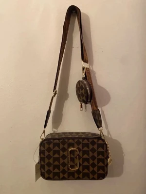 Brun axelväska med geometriskt mönster - Snygg brun axelväska med geometriskt rutigt mönster och guldfärgade detaljer. Väskan har bred axelrem med logga och en liten rund myntväska på remmen. Stora guldfärgade dragkedjor och dekorativa hängen ger extra stilpoäng.