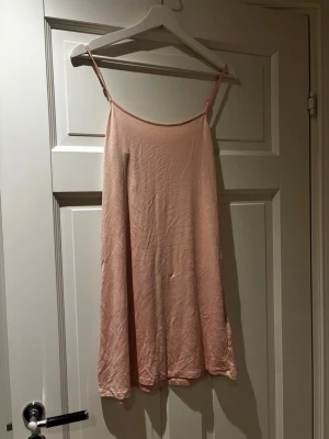 Ljusrosa ärmlös kortklänning - Säljer en enkel och stilren ljusrosa klänning med smala axelband och rak passform. Klänningen är kort och har en mjuk, slät yta som känns skön mot huden. Perfekt för varma dagar och lätt att matcha med accessoarer. Kan användas som den är eller som underklänning