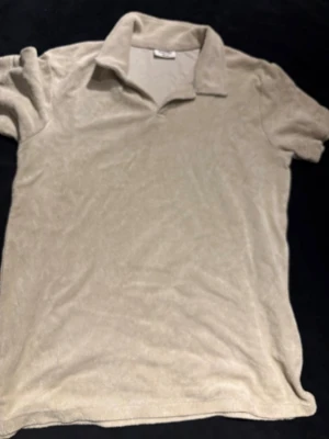 Beige frotté polotopp från Kappahl - Säljer en beige polotopp från Kappahl i mjuk frotté. Toppen har klassisk krage och korta ärmar, perfekt för chill dagar. Materialet är skönt mot huden och ger en avslappnad vibe. Passar dig som gillar neutrala färger och enkel stil.