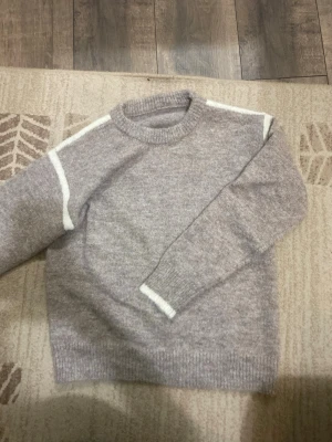 Beige stickad tröja med vita detaljer - Mysig beige stickad tröja med vita ränder längs axlar och ärmslut. Rund hals och ribbade muddar ger en chill och avslappnad vibe. Perfekt för lager-på-lager under kyliga dagar. Passformen är normal och tröjan har långa ärmar. tröjan är använde endast 1 gång. Vet tyvärr inte vart den är ifrån då det inte finns nån lap😊