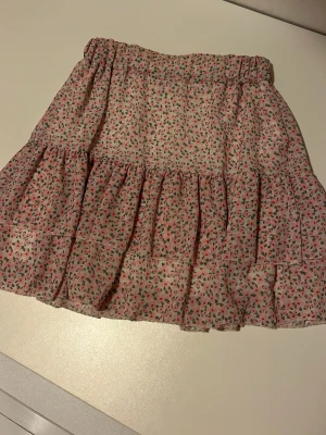 Blommig volangkjol från Holly's - Supersöt kort kjol från Holly's i storlek S med blommigt mönster i rosa och grönt. Kjolen har volanger och resår i midja som ger en skön passform. Materialet är lätt och luftigt, perfekt för varma dagar. ”Underkjolen” har rispat upp sig lite men inget som förstör eller syns!