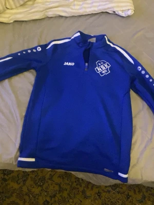 Blå JAKO NSK träningströja 164 - Säljer en blå träningströja från JAKO med NSK-logga och texten Nykvarns SK på ryggen. Tröjan har vit detalj på ärmar och nederkant samt halv dragkedja framtill. Perfekt för träning eller uppvärmning.