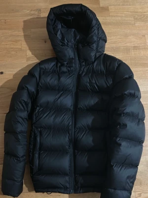 Everest M Expedition Down Jacket  - Svart dunjacka från Everest med stor, mysig huva och dragkedja framtill. Jackan har puffad design och är tillverkad i syntetmaterial med dunfyllning. Perfekt för kalla dagar och har en clean look som funkar till det mesta men reflexen inuti fickan saknas och det är den enda saken som inte finns. Ny pris är 2500kr priset kan diskuteras. Den är också sällsynt använd har bara varit i garderoben för det mesta.