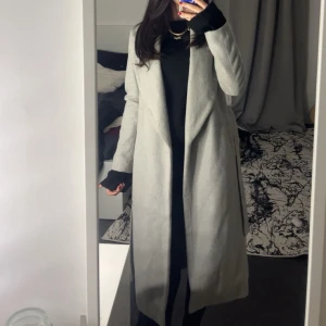 Ljusgrå lång kappa  - Säljer en stilren ljusgrå kappa med lång passform och klassisk krage. Jackan har en enkel och clean look utan synliga knappar eller dragkedja, perfekt för dig som gillar minimalistisk stil. 