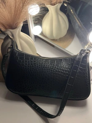 Svart axelväska i croco-mönstrat skinn - Säljer en svart axelväska med croco-mönstrad yta och guldfärgade detaljer vid dragkedjan. Väskan har en stilren, avlång form och justerbar rem som gör den enkel att slänga över axeln. Perfekt för dig som gillar trendiga accessoarer med lite edge.