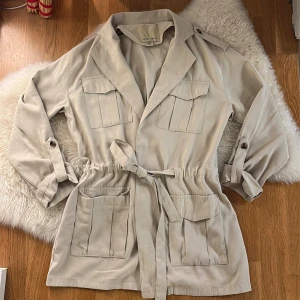 Beige Trenchcoat från Amisu med bälte - Snygg beige parkajacka från Amisu i storlek 34. Jackan har fyra stora fickor framtill, knytbälte i midjan och knappar vid ärmsluten. Perfekt för dig som gillar en chill och stilren look. Passar bra till vår och höst.