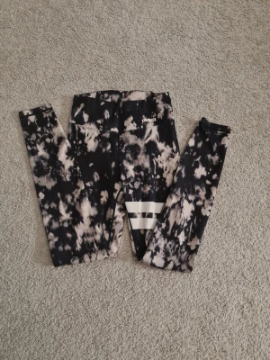 Svarta och vita tights från Stronger - Supersnygga tights från Stronger i svart och vitt batikmönster. Hög midja och två vita ränder på ena benet ger en sportig vibe. Tillverkade i återvunnen polyester och elastan för skön stretch. Perfekta för träning eller chill.