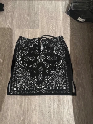 Snygg kort kjol - S - Säljer en svart kortkjol från Fasister med vitt bandanamönster och snörning framtill. Kjolen har slits på sidorna och är i mjukt material som känns skönt mot huden. Perfekt för dig som vill ha en edgy och trendig look.