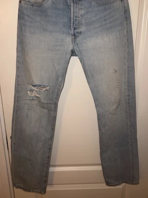 Levi's 501 ljusblå jeans - Ljusblå Levi's 501 jeans med klassisk rak passform och femficksdesign. Jeansen är tillverkade i slitstarkt bomullsdenim och har en snygg vintagekänsla med lätt slitning på benen. Perfekta för dig som gillar tidlös stil och vill ha ett par ikoniska jeans.