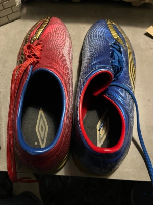Umbro fotbollsskor i rött och blått - Säljer ett par Umbro fotbollsskor med coolt mönster i rött och blått, med detaljer i guld och svart. Skorna har snörning och en platt sula med bra grepp. Materialet är syntet och designen har vita linjer som ger en futuristisk känsla. Perfekt för dig som vill sticka ut på planen.