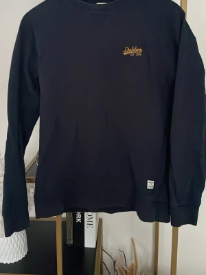 Mörkblå sweatshirt från Dobber - Snygg mörkblå sweatshirt från Dobber med gul logotyp på bröstet och liten vit etikett vid nederkanten. Tröjan har rund hals och långa ärmar, perfekt för chill dagar. Materialet är mjuk bomull som känns skönt mot huden.