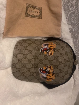 Keps G - Säljer en snygg G keps med klassiskt GG-mönster i beige och brunt. Framsidan har två broderade tigrar i orange, svart och vitt. Baksidan är i svart mesh och justerbar med grönt och rött band. Perfekt för dig som vill sticka ut med lyxig streetstyle. Påser ingår 