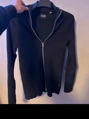 Svart kofta från Only & Sons - Svart ribbstickad kofta från Only & Sons med hel dragkedja framtill. Långärmad modell med stilren design och hög krage. Perfekt att slänga på sig när det är lite kyligare ute. Materialet är mjukt och känns skönt mot huden.