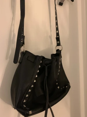 Svart axelväska med nitar  - Cool svart axelväska i skinnimitation med silverfärgade nitar längs kanterna och justerbar axelrem. Väskan har snörning upptill för stängning samt en magnet! Bohochic inspiration 