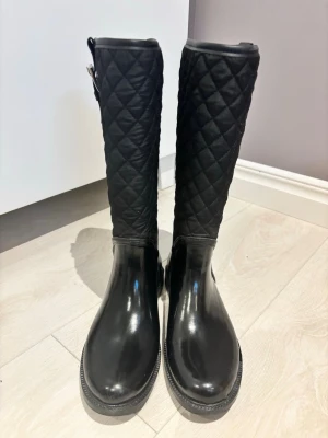 Svarta quiltade stövlar med spänne - Säljer ett par svarta stövlar med quiltad textil upptill och glansig syntet nedtill. De har ett snyggt spänne i silver vid skaftet och rund tå. Perfekta för dig som vill ha en stilren look med lite edge.