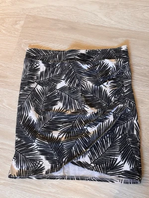 Svartvit omlottkjol med palmblad - Kort kjol med svartvita palmbladsmönster och omlottdetalj. Kjolen har en skön passform och är perfekt för varma dagar. Tillverkad i mjukt material som känns bekvämt mot huden.