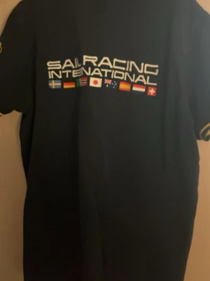 Svart Sail Racing polotopp med flaggor - Svart polotopp från Sail Racing med internationella flaggor och text på ryggen. Framsidan har svensk flagga och logga, samt krage och knappar. Ärmarna har gula detaljer och tryck. Perfekt för dig som gillar segling och sportig stil.