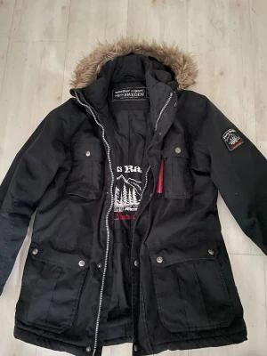 Svart parkajacka från Weather Report - Parkajacka i svart med fuskpäls på huvan och broderad Parks Ranger-logga på insidan. Jackan har flera fickor med knappar, dragkedja framtill och patch på ärmen. Perfekt för kalla dagar och outdoor-vibes I mycket fint skick knappt använd 