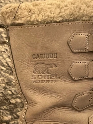 Sorel Caribou vinterstövlar strl 38 - Säljer ett par Sorel Caribou vinterstövlar i beige och brunt med varm ullfoder och snörning framtill. Stövlarna är vattentäta och har en robust platt sula som ger bra grepp. Perfekta för kalla dagar och snö. Fodret är fluffigt och skönt, och utsidan är i slitstarkt skinn.