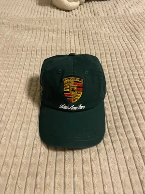 Aimé Leon Dore x Porsche Keps - Mörkgrön keps från Aimé Leon Dore och Porsche med broderad Porsche-logga framtill och gul text på sidan. Justerbart spänne bak i mässing med ingraverad blomdetalj. Kepsen är i bomull och har klassisk form med böjd skärm.