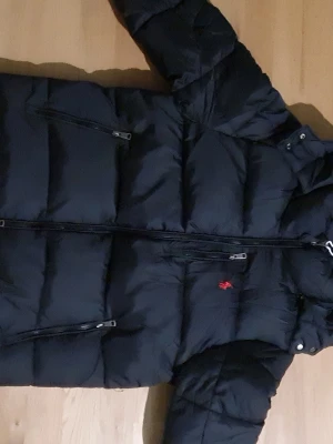 Svart pufferjacka från Polo Ralph Lauren - Svart pufferjacka från Polo Ralph Lauren med röd broderad logga på bröstet. Jackan har flera dragkedjeförsedda fickor och hög krage. Materialet är polyester och jackan har en normal passform. Perfekt för kalla dagar med sin tjocka och quiltade design.