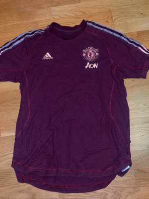 Man Utd tröja - Bilden visar ingen sko, utan en vinröd Manchester United t-shirt från Adidas med ljusrosa detaljer och klubbmärke på bröstet.