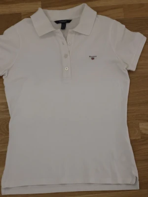 Vit pikéblus från GANT XS - Säljer en klassisk vit pikéblus från GANT i storlek XS. Toppen har korta ärmar, krage och knappar framtill. Materialet är mjuk bomull och den har en liten broderad GANT-logga på bröstet. Perfekt för en clean och sportig look.