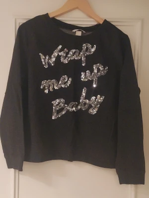Svart tröja med paljetter från H&M - Cool svart tröja från H&M i storlek M med texten 'Wrap me up Baby' i glittriga silverpaljetter på framsidan. Tröjan har rund hals och långa ärmar, perfekt för dig som gillar att sticka ut med lite extra bling.