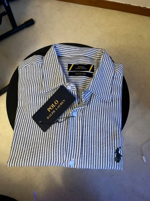 Randig skjorta från Ralph Lauren - Snygg skjorta från Ralph Lauren i storlek medium. Skjortan har ett klassiskt krage, svarta och vita vertikala ränder, knappar framtill och den ikoniska broderade loggan på bröstet. Perfekt för dig som gillar stilrena och tidlösa plagg.