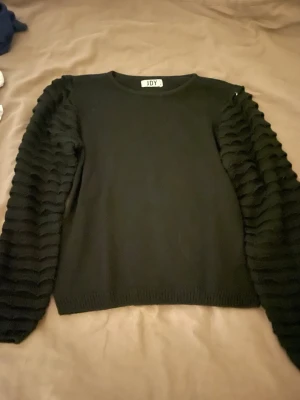 Svart stickad tröja JDY med volymärmar - Svart stickad tröja från JDY i storlek S. Tröjan har unika volymärmar med vågigt, ribbat mönster som ger en cool och trendig look. Rund halsringning och ribbade muddar vid ärmslut och nederkant. Perfekt för dig som gillar detaljer och vill sticka ut.