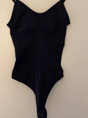 Svart bodysuit med justerbara band - Svart bodysuit med smala axelband och formade kupor. Bodyn har justerbara axelband och knäppning med hyskor i grenen. Materialet är stretchigt och följsamt, perfekt för en stilren look.