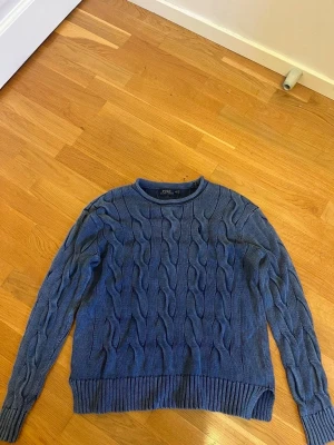 Blå kabelstickad tröja Polo Ralph Lauren - Kabelstickad blå tröja från Polo Ralph Lauren i storlek M. Tröjan har rund halsringning, ribbade muddar och nederkant samt ett klassiskt kabelmönster. Perfekt för lager-på-lager under kyliga dagar.