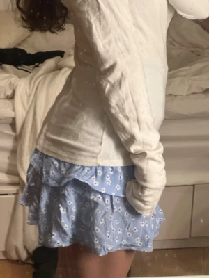 Blå blommig volangkjol  - Supersöt blå kortkjol med vita små blommor och volangdetaljer. Kjolen har en smockad midja som gör den bekväm och sitter snyggt. Perfekt för dig som gillar en feminin och fräsch stil. Passar till både sneakers och sandaler.