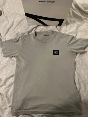  Stone Island t-shirt junior  - Säljer en grå t-shirt från Stone Island med klassisk svart patch på bröstet. T-shirten har rund hals och korta ärmar, Perfekt för dig som gillar stilrena och ikoniska plagg. Passar folk som är runt 150cm säljer pågrund av för liten, köpt på NK i Göteborg mer bilder kan fixas släng bara förbi ett sms vid minsta funderingar 
