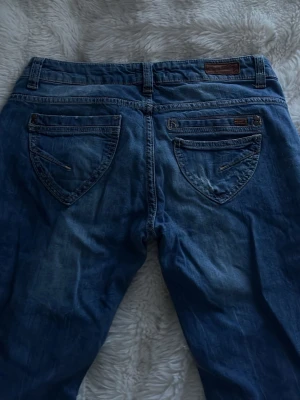 Blå raka jeans från Fuga, storlek 34 - Säljer ett par blå jeans från Fuga i rak modell med slitna detaljer och snygg tvätt. Jeansen har klassiska fem fickor, dragkedja och knapp framtill samt coola sömmar och logga på fickan. Perfekta för en avslappnad look.