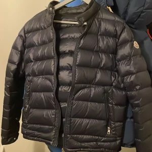 Mörkblå Moncler Acorus - Säljer en mörkblå pufferjacka från Moncler med klassisk logga på ärmen. Jackan har hög krage, dragkedja framtill och två sidofickor med dragkedja. Materialet är glansigt och lätt, perfekt för kyliga dagar. Skicka pm vid frågor. 