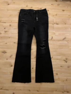 Svarta bootcut jeans från Fashion Nova - Säljer ett par svarta bootcut jeans från Fashion Nova med slitna detaljer och råa kanter. Jeansen har en klassisk femficksdesign och coola slitningar på både fram- och baksida. Materialet är stretchigt denim som sitter snyggt och bekvämt på kroppen.